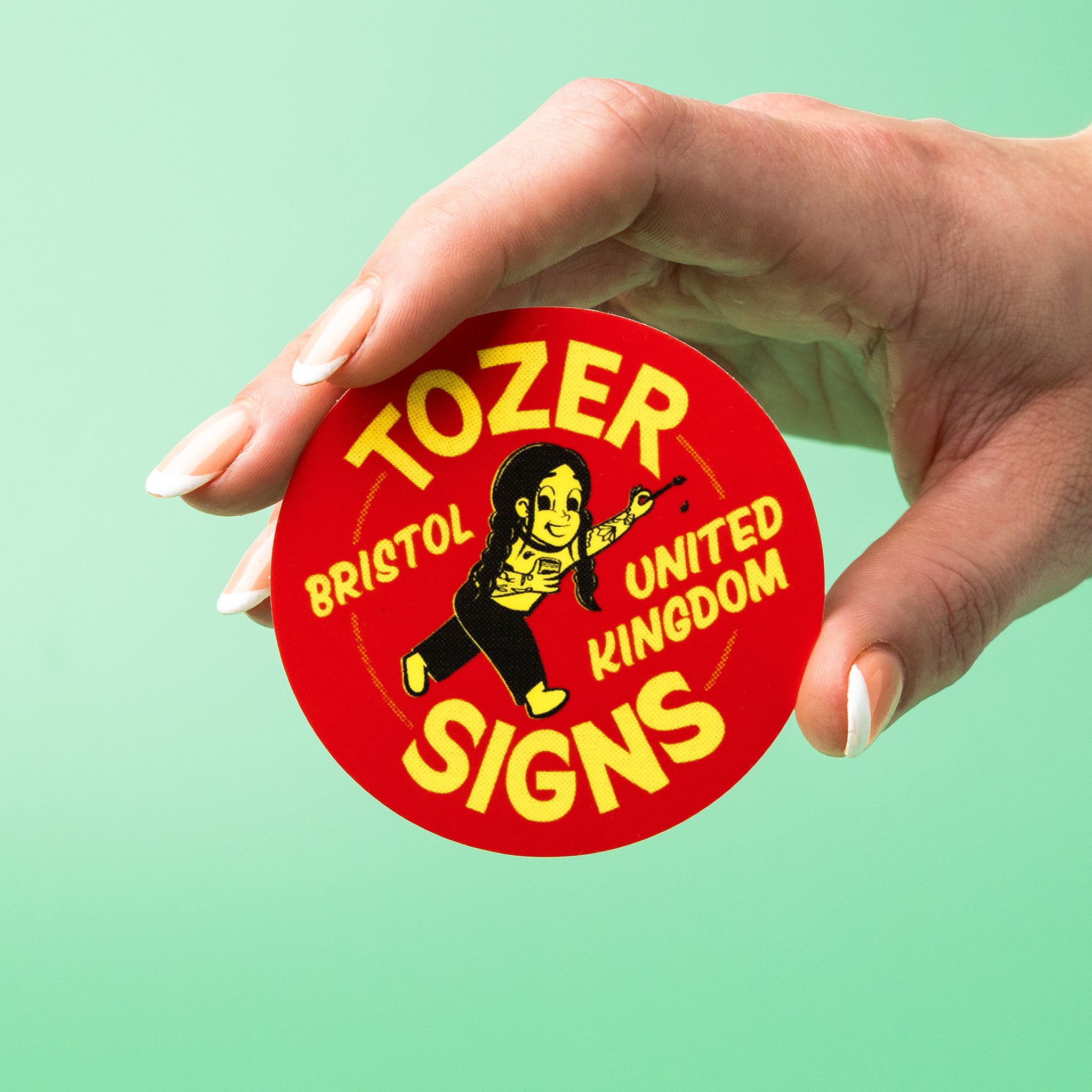 Hand holding a red and yellow 'Tozer Bristol United Kingdom Signs' custom vinyl sticker against a light green background.
