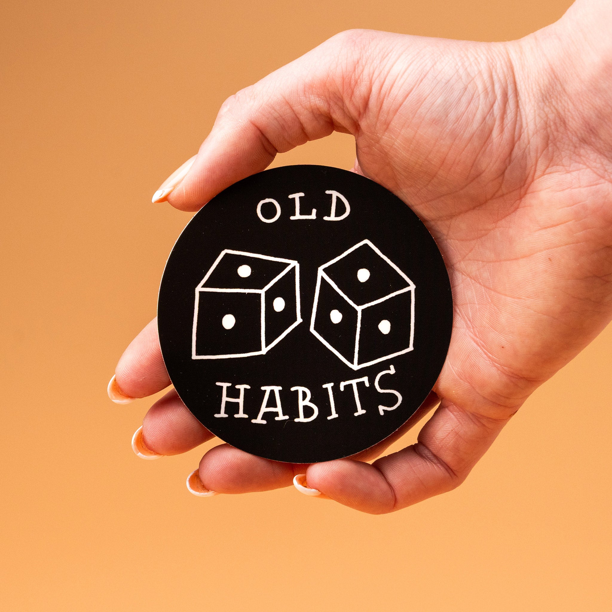 Hand holding a black round custom vinyl sticker with dice graphics and 'Old Habits' text on an orange background.