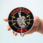 Circular custom vinyl sticker with 'Grim Team Paris' design held by a hand against a light blue background.