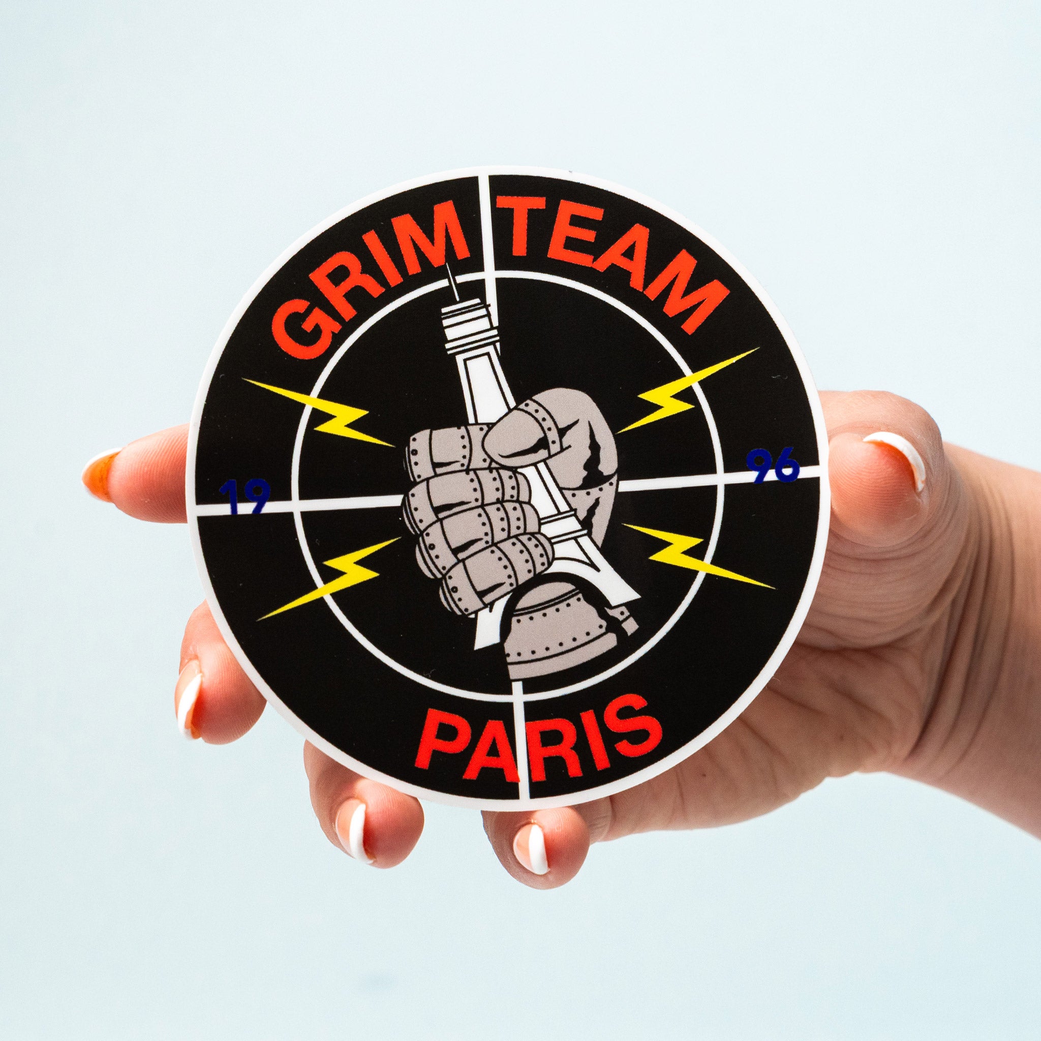 Circular custom vinyl sticker with 'Grim Team Paris' design held by a hand against a light blue background.