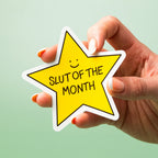 Hand holding a yellow star-shaped custom vinyl sticker with 'Slut of the Month' text on a light green background.