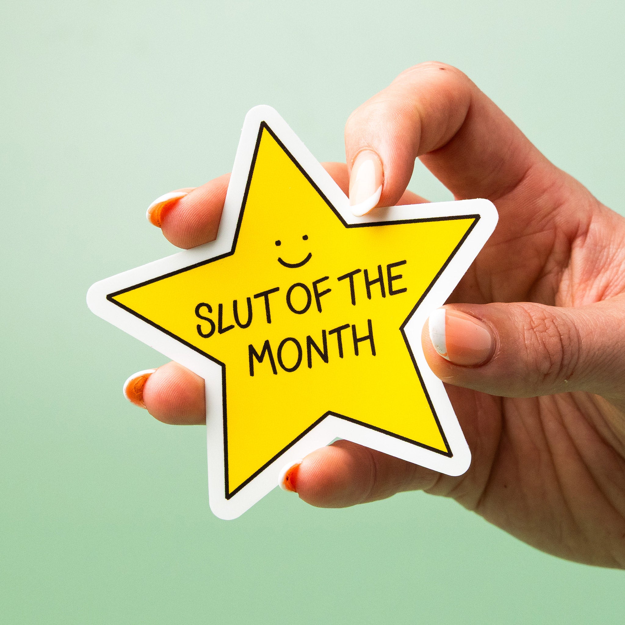 Hand holding a yellow star-shaped custom vinyl sticker with 'Slut of the Month' text on a light green background.
