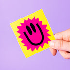 Hand holding a yellow custom vinyl sticker with a pink smiley face design against a purple background.