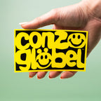 Hand holding a yellow and black custom vinyl sticker with 'conzoglobel' text and smiley face design on a light green background.